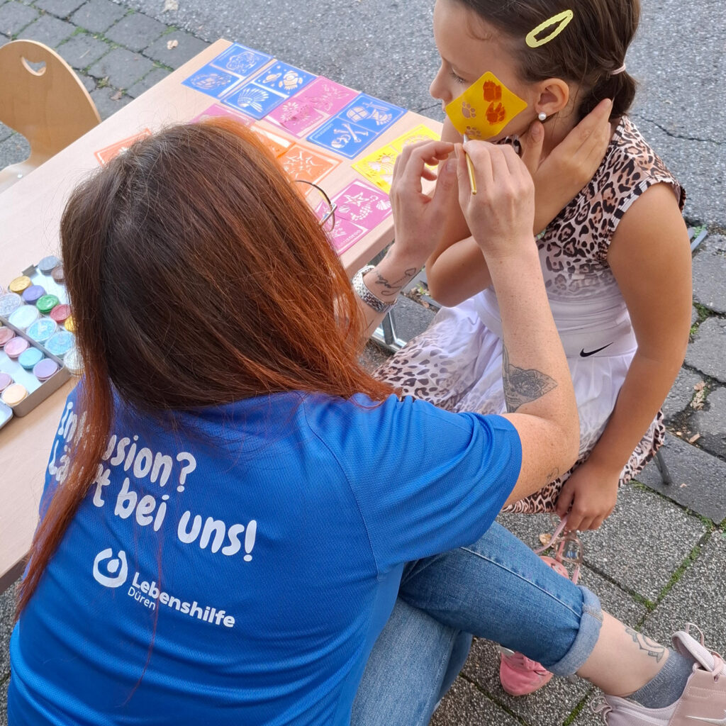Eine Mitarbeiterin der Lebenshilfe schminkt ein Kind beim Ortsfest in Merzenich